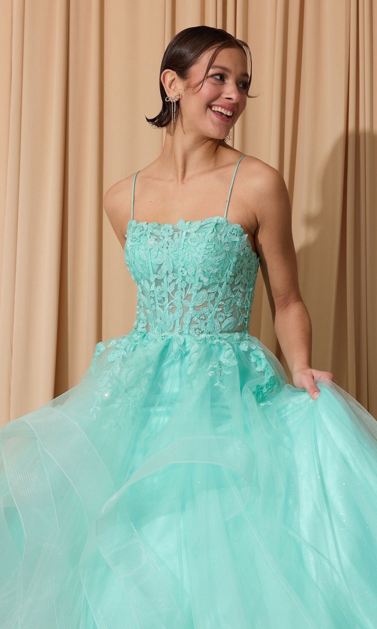 A woman in the Eureka EK167 long prom dress with a light blue glitter tulle skirt, floral lace corset bodice, and thin straps stands smiling in front of beige curtains, holding her dress.